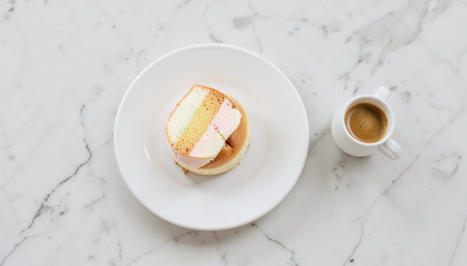 Overhead flat-lay photograph of a pastel de nata on a white saucer next to a small espresso cup on a marble table, cream and brown tones, soft morning light.
